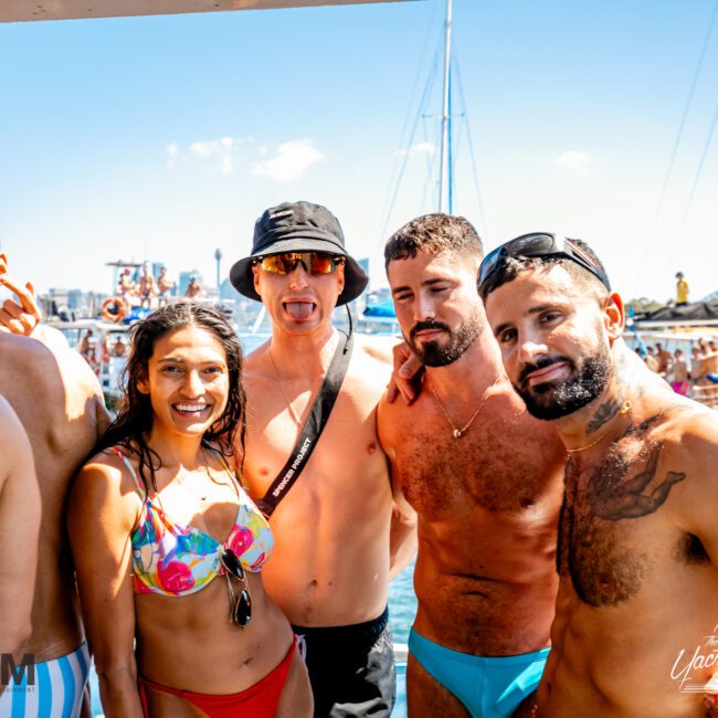 A group of people in swimsuits is gathered together on a boat under clear blue skies, smiling and enjoying a sunny day on the water. The background features other boats and what seems to be a cityscape with a bridge, typical of Boat Parties Sydney The Yacht Social Club events.