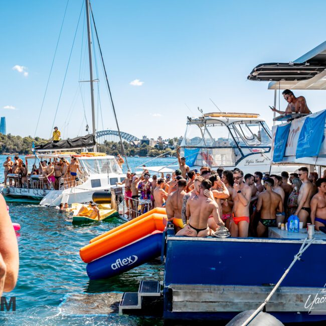 A lively party scene on the water with numerous people on boats and inflatable rafts. The boats, including luxury yachts from Sydney Harbour Boat Hire, are docked close together. Many attendees are wearing swimwear, enjoying the sun. The background features city buildings and a bridge.