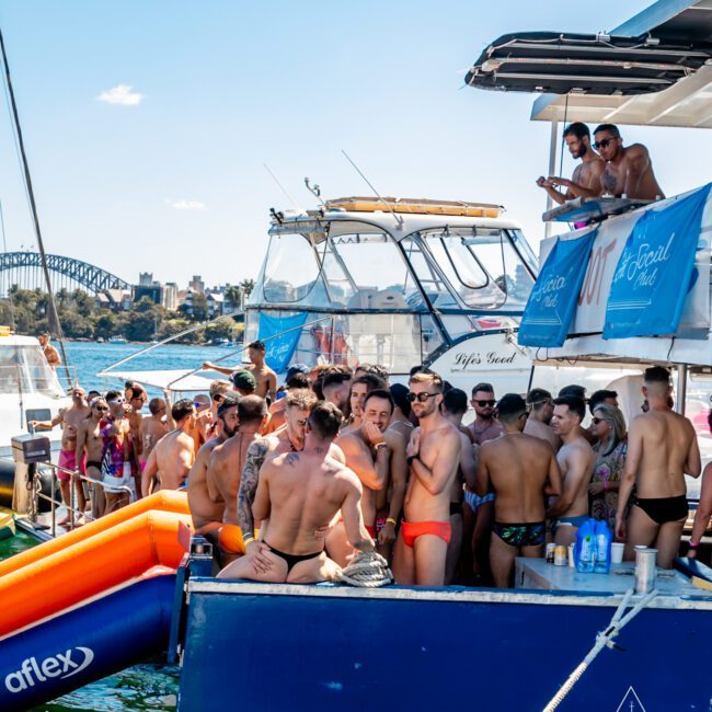 A lively yacht party with a large group of people in swimwear enjoying the sun. Many are gathered on the deck near inflatables, with some on an upper level. A city skyline and a large bridge are visible in the background, courtesy of Sydney Harbour Boat Hire The Yacht Social Club.