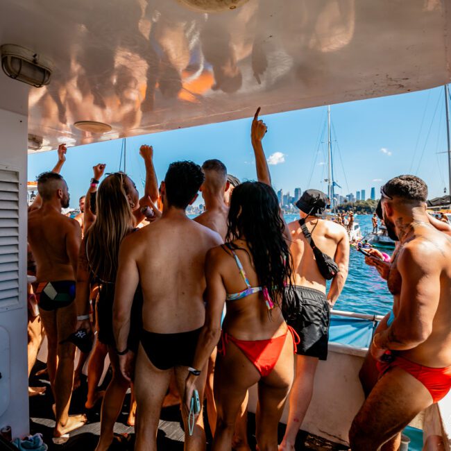 A group of people in swimwear stands on a boat deck, facing away from the camera and raising their arms. They appear to be enjoying a lively atmosphere under the bright sun. Other boats and a city skyline can be seen in the background, typical of Boat Parties Sydney with The Yacht Social Club.