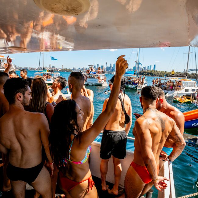 A group of people on a boat partying in swimwear, with some dancing and others socializing. In the background, several boats and people can be seen on the water. The scene is lively, with clear skies and a city skyline visible in the distance. It's an iconic Boat Party hosted by The Yacht Social Club in Sydney.