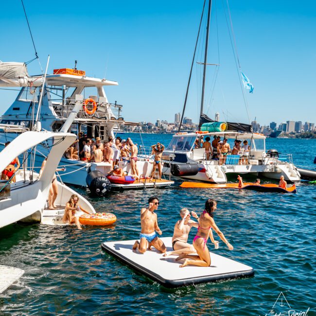 A lively scene on the water shows several boats gathered together with people socializing, swimming, and sunbathing. Three individuals are seen on an inflatable platform enjoying the sun. The backdrop includes a city skyline under a clear blue sky, perfect for events like The Yacht Social Club Event Boat Charters.