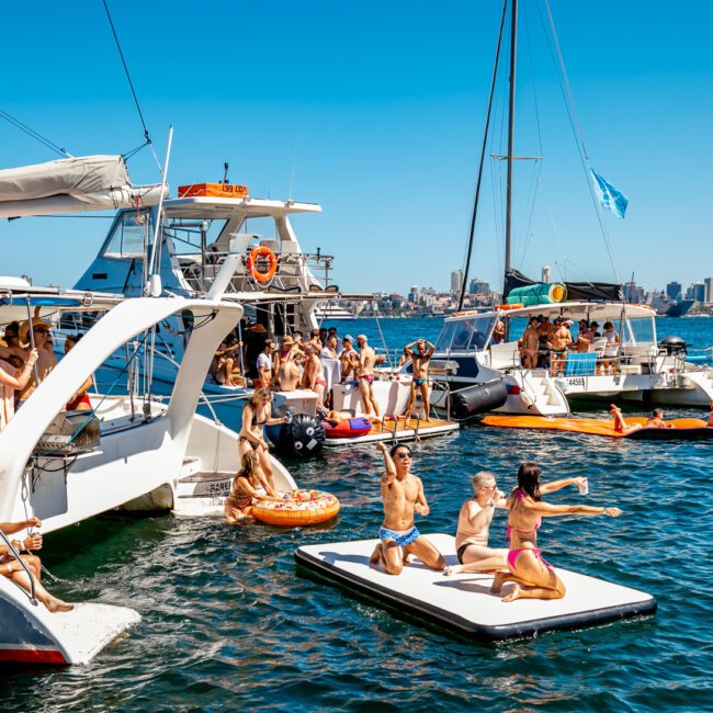 People on boats at a lively marina party on a sunny day. Some are swimming, lounging on inflatable platforms, and dancing. The scene includes multiple boats, clear blue water, and a background of city skyscrapers. The overall atmosphere is festive and vibrant with Luxury Yacht Rentals Sydney.