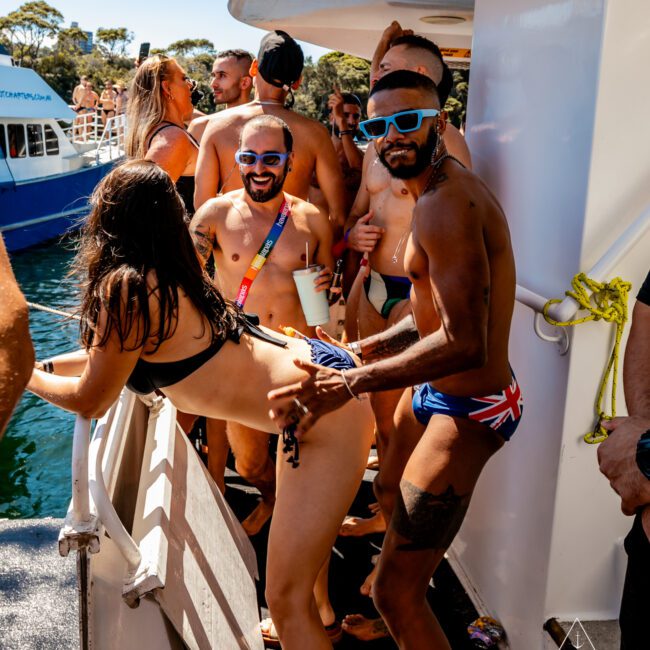 A group of people in swimwear dancing and enjoying themselves on a luxury yacht. The man in the foreground is in blue swim trunks, wearing sunglasses, and has his hand on the back of a dancing woman in a bikini. Other people are smiling and socializing, enjoying what feels like one of the best Boat Parties Sydney has to offer.