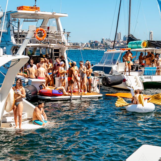 A lively scene of people enjoying a sunny day on a boat and in the water. Several individuals are lounging on the deck, swimming, and using inflatables, including a large pelican float. In the background, a city skyline is visible against a clear blue sky—perfect for Luxury Yacht Rentals Sydney.