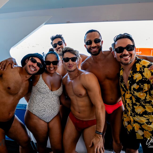 A group of six people, four men and two women, smiling and posing together on a boat from The Yacht Social Club Sydney Boat Hire. The men are wearing swim trunks and sunglasses, while the women are in swimsuits and sunglasses. An inflatable raft is visible in the background.