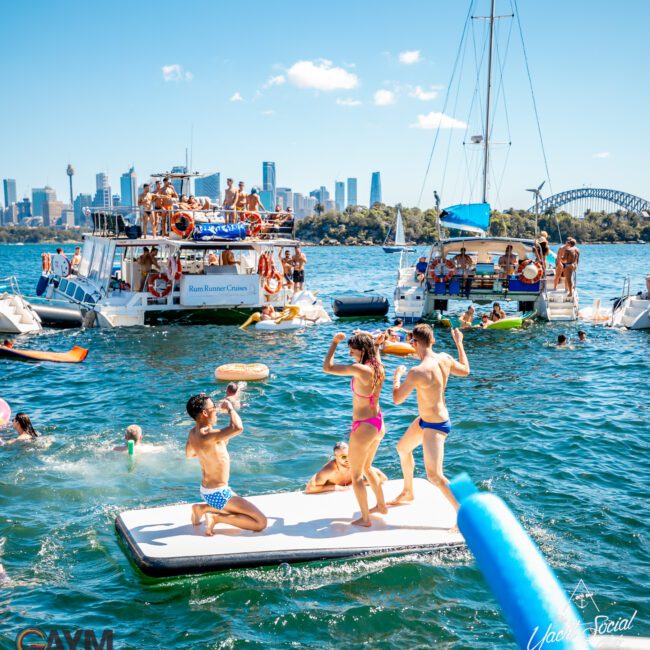 People are enjoying a sunny day on the water with boats and inflatables. Individuals are seen swimming, playing, and relaxing against a backdrop of a city skyline and a bridge. The atmosphere is lively with a clear blue sky, perfect for Luxury Yacht Rentals Sydney from The Yacht Social Club.