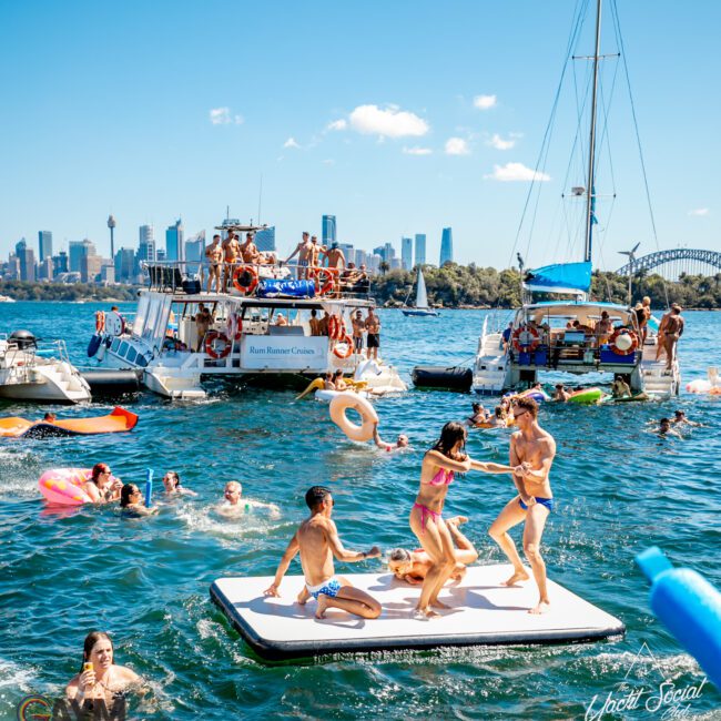 A lively scene of people enjoying a sunny day on the water. Several boats are anchored close to each other with people swimming, playing on a floating mat, and relaxing on inflatables. A city's skyline and a bridge are visible in the background. This could easily be part of The Yacht Social Club Event Boat Charters experience.