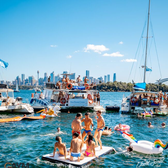 A vibrant scene on the water shows people enjoying a sunny day on boats and inflatables. Some are playing on a floating platform, while others relax on various watercraft. The Yacht Social Club Event Boat Charters offer an exciting experience, with the city skyline visible in the background under a clear, blue sky.