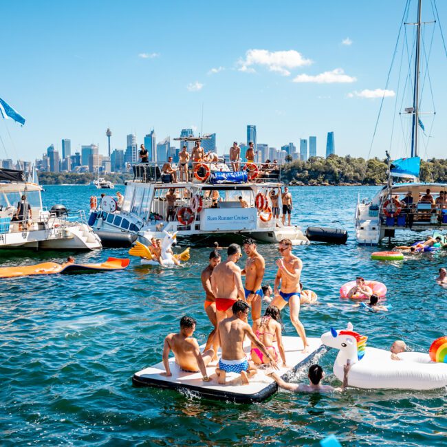 A lively scene of people enjoying a sunny day on Sydney Harbour, often facilitated by The Yacht Social Club Sydney Boat Hire. Multiple boats are anchored, with people swimming and lounging on inflatable rafts, including a unicorn float. The Sydney skyline and Harbour Bridge provide a stunning backdrop.