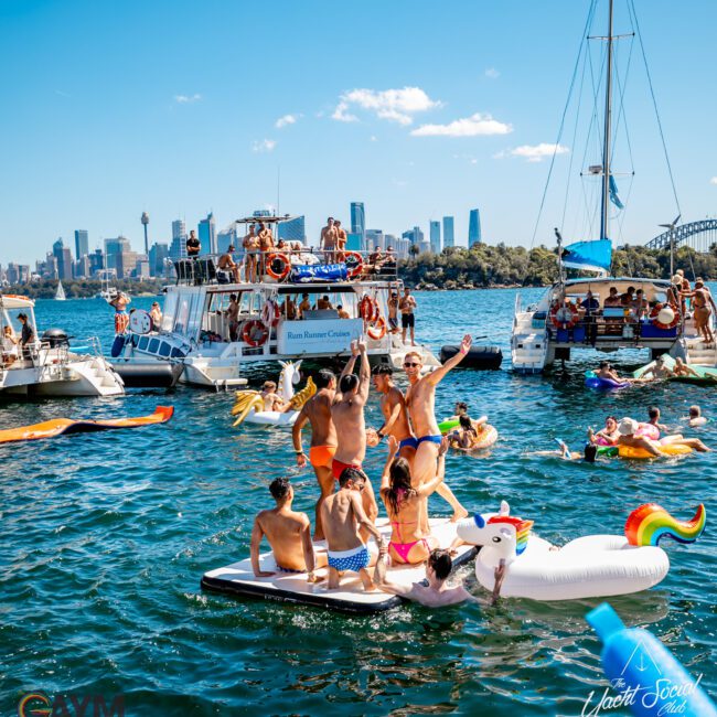 People enjoying a sunny day on a crowded waterway with boats and inflatables. Some are on a large unicorn float, while others are on boats or swimming. The background features a skyline with high-rise buildings and greenery, reminiscent of Boat Parties Sydney The Yacht Social Club.