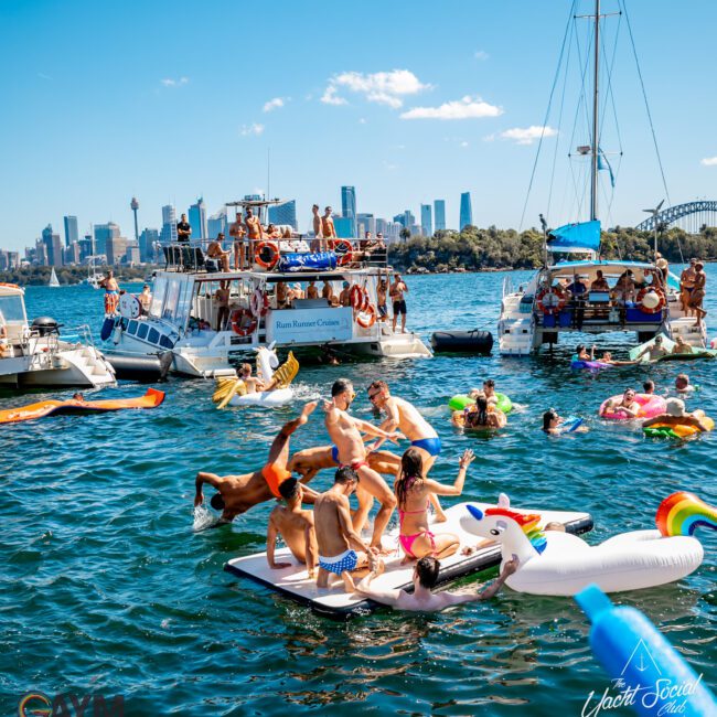 A lively scene on a sunny day where people are swimming and lounging on inflatables near anchored boats. Hosted by The Yacht Social Club Sydney Boat Hire, the background features a city skyline and a blue sky. The water is filled with colorful floats, including a large unicorn inflatable.