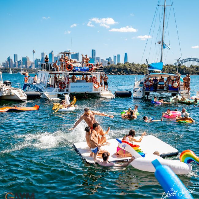 A lively gathering on a sunny day, with people enjoying the water on colorful inflatables near several boats. The city skyline is visible in the background. Some individuals relax on a floating platform, while others swim and socialize in the water at The Yacht Social Club Event Boat Charters.