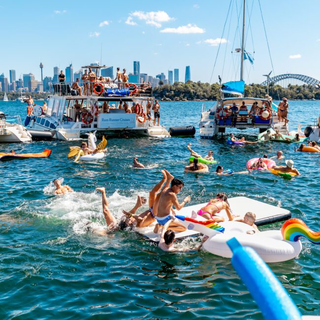 A vibrant scene of people enjoying a sunny day on the water near a city skyline. Several boats are anchored, with people swimming, lounging on inflatables, and socializing. The iconic Sydney Harbour Bridge is visible in the background, highlighting the fun of The Yacht Social Club Event Boat Charters.