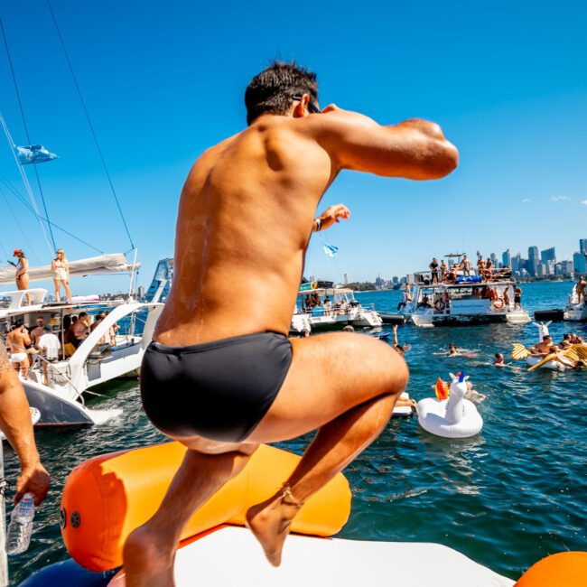 A muscular man in swim trunks jumps into the water from a yacht while another man stands nearby holding a drink. Several boats and yachts are anchored in the background with people socializing and swimming on a sunny day. The city skyline is visible in the distance, indicating a vibrant Boat Parties Sydney The Yacht Social Club event.