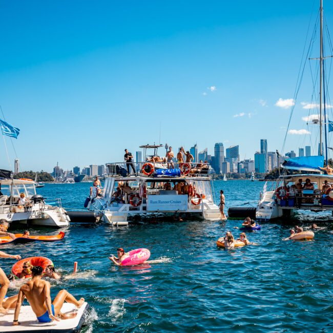 A vibrant scene on a sunny day with multiple boats anchored close together on the water, people swimming, floating on inflatables, and relaxing on deck. The city skyline is visible in the background, clear blue skies, and a festive atmosphere with music and laughter—truly a perfect day for Luxury Yacht Rentals Sydney.