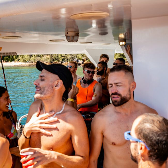 A group of shirtless people in swimwear enjoy a boat party hosted by The Yacht Social Club Sydney Boat Hire. The group is gathered under a shaded area, some wearing sunglasses and caps. One person is holding a red cup. The background shows a body of water and a rocky shoreline with trees.