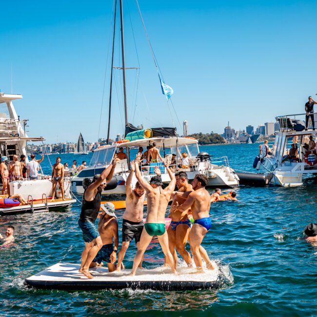 A group of people enjoy a sunny day on the water, with multiple boats docked nearby. Some individuals dance and cheer on a floating platform in the center, while others relax and socialize on the boats. The city skyline is visible in the background during The Yacht Social Club Event Boat Charters.