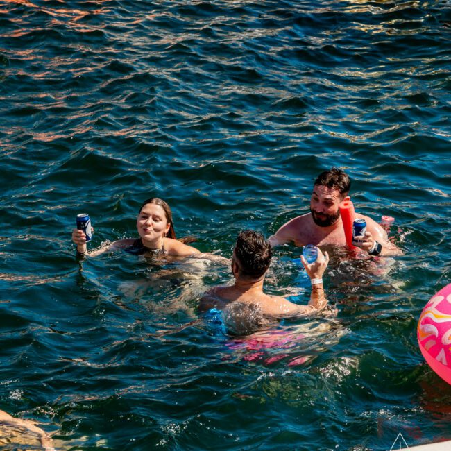 Three people are enjoying themselves in the water, holding drinks and smiling. They are surrounded by inflatables, including a visible white and gold inflatable in the top right corner. The water is dark blue and rippled. Logos of "The Yacht Social Club" and "Night Swim Club" are visible.