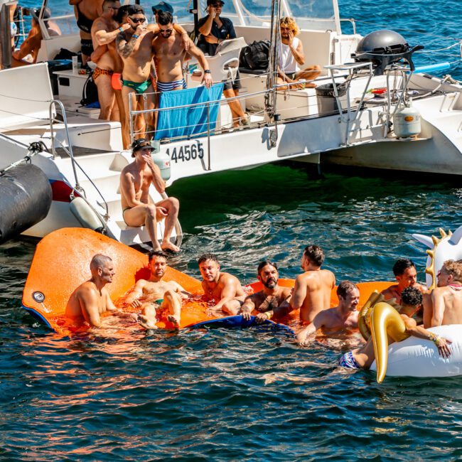 A group of people enjoying time in the water next to a docked boat. Some are on inflatable floats, including an orange octopus and a white unicorn with gold wings. Others are swimming or sitting on the boat. The day is sunny, and the atmosphere at The Yacht Social Club Sydney Boat Hire is lively and festive.