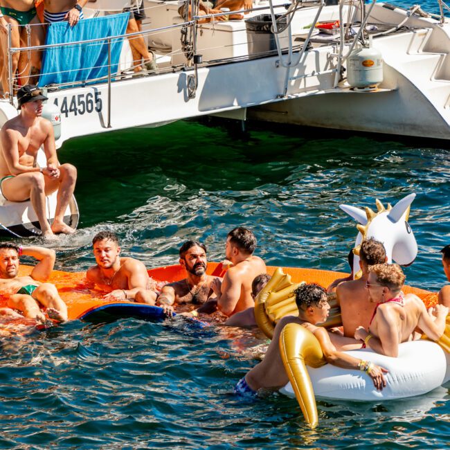 A group of people enjoying a sunny day on the water near a boat. Some are sitting on a large floating mat, others on inflatable pool toys, including a unicorn. They appear to be relaxing and socializing, surrounded by clear blue water as part of The Yacht Social Club Event Boat Charters.