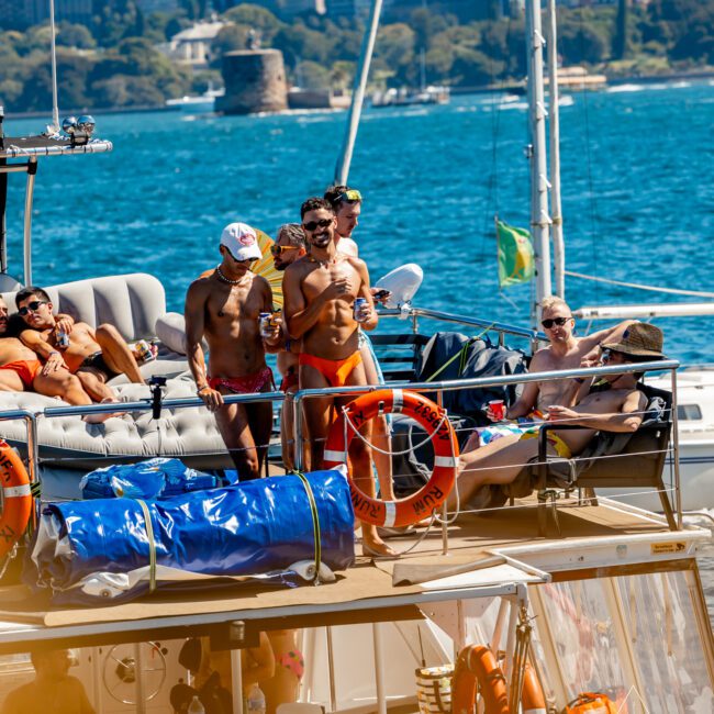 A group of people in swimwear socialize on a boat deck under bright sunlight. Some sit and lounge while others stand near railings. The background features a scenic view of a city skyline, blue water, and trees. The atmosphere is lively and relaxed at Boat Parties Sydney hosted by The Yacht Social Club.