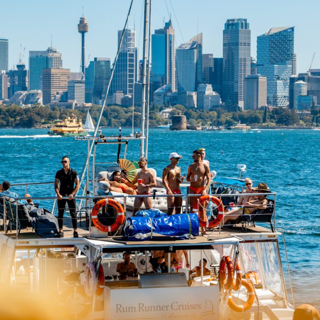 A group of people enjoy a sunny day on a boat named "Rum Runner Cruises" in the bustling Sydney Harbour with a cityscape in the background. The boat is equipped with life preservers and various boating gear. The city skyline features tall buildings and a prominent tower, capturing the essence of Luxury Yacht Rentals Sydney.