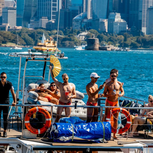 A group of men in swimwear enjoy a sunny day on a yacht, some relaxing and others standing. Surrounded by water with a city skyline in the background, they embrace the vibes of The Yacht Social Club Sydney Boat Hire. The yacht features visible life rings and a wrapped package on the deck.