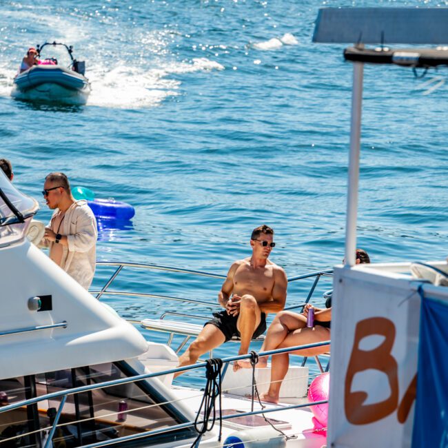 A shirtless man relaxes on the bow of a yacht in sunny weather, courtesy of Luxury Yacht Rentals Sydney. Several other individuals are on board as well. In the background, a small boat speeds across the water with a wake trailing behind it. "AYM" and "Yacht Social" logos are visible on the image.