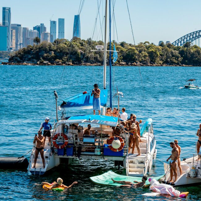 A vibrant scene of two docked yachts in a sunny harbor filled with people enjoying water activities. Individuals are swimming, lounging on inflatables, and socializing on the boats, reminiscent of The Yacht Social Club Sydney Boat Hire events. The city skyline and a large arch bridge are visible in the background.