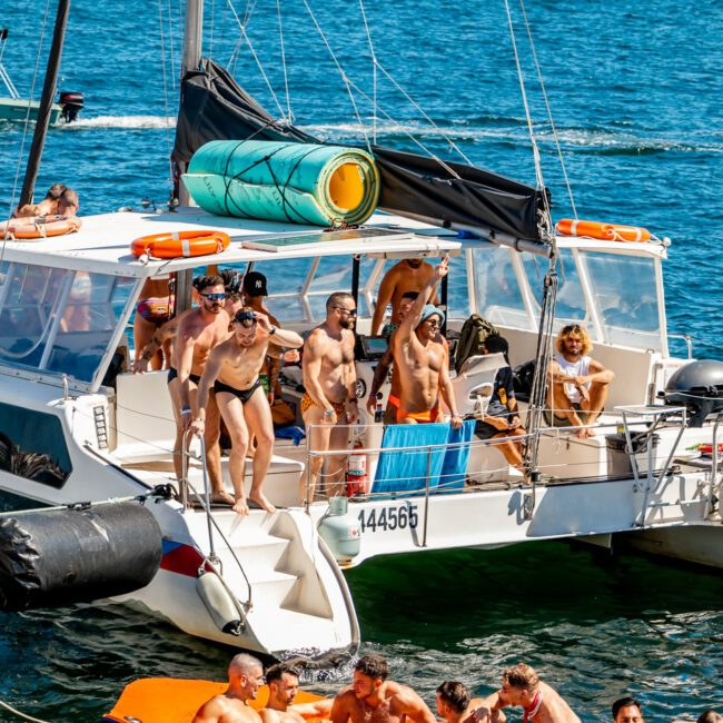 A lively group of people enjoying a sunny day on and around a catamaran. Some are on the boat, while others play in the water on inflatable floats, including a unicorn float. The backdrop includes Sydney Harbour with its cityscape and calm blue waters, perfect for Boat Parties Sydney The Yacht Social Club.