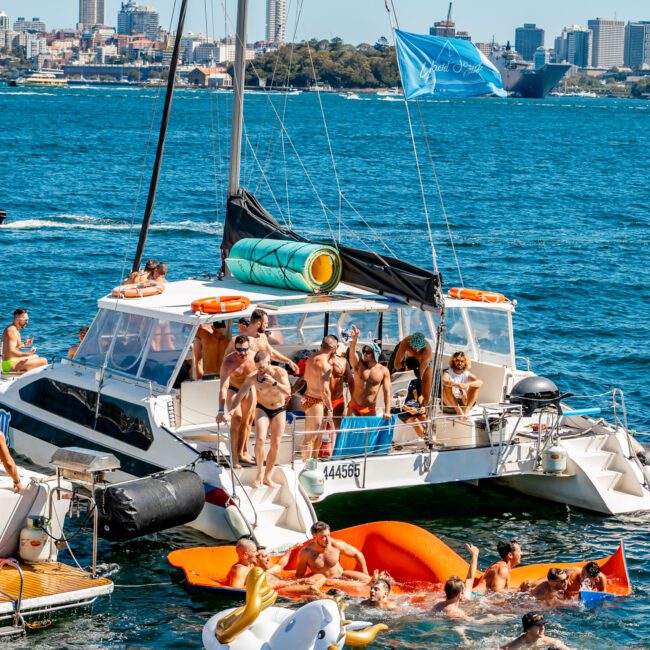 A group of people are enjoying a sunny day on a catamaran and in the water around it. Some are lounging on the boat while others swim and play with inflatable floats. The backdrop features an urban skyline and a calm blue sea, showcasing the vibrant atmosphere of Boat Parties Sydney by The Yacht Social Club.