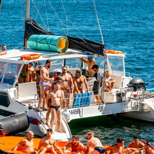 A group of people on a luxury yacht enjoying a sunny day. The boat has a blue flag and is anchored on a clear blue lake, with people lounging on the deck and swimming nearby. City buildings are visible in the background. Various inflatables are in the water, making it an ideal scene for Boat Rental and Parties Sydney The Yacht Social Club.