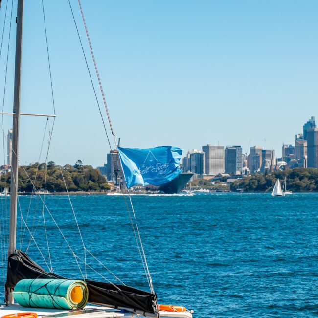 A sailboat on calm blue waters with city skyscrapers in the background. The boat has a visible mast and rolled-up sail. Other boats and a small island can be seen along the waterfront under a bright, clear sky. Perfect for Sydney Harbour Boat Hire with The Yacht Social Club's event charters.