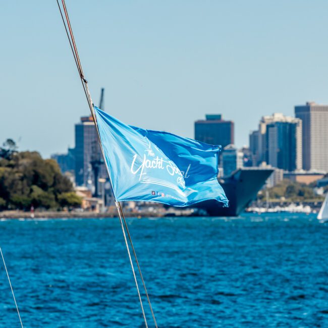 A blue flag with white text reading "The Yacht Social Club" flutters in the wind on a sailboat. In the background, calm blue waters showcase other sailboats, and a distant city skyline is visible under a clear, sunny sky. Trees line the left shoreline.