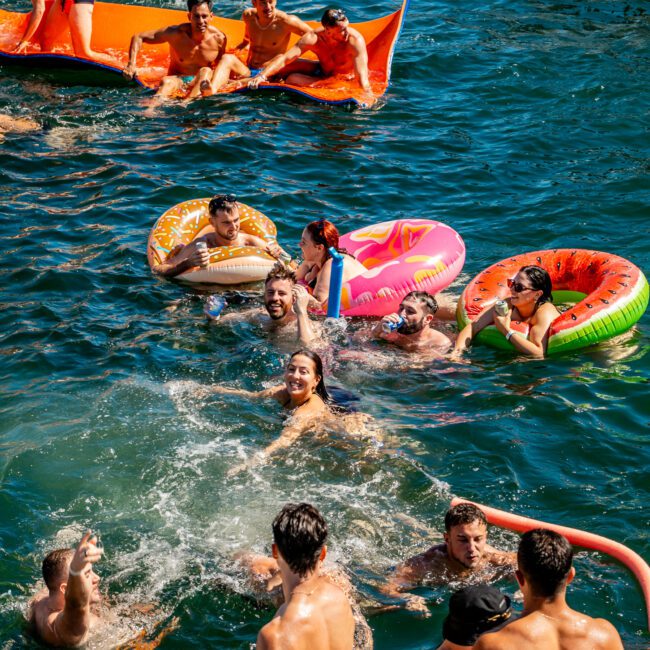 A group of people enjoys swimming and floating on inflatable tubes in the water on a sunny day. Some are on a large orange float, while others are in the water or on colorful donut-shaped inflatables. The Yacht Social Club Event Boat Charters adds to the lively atmosphere filled with fun and laughter.