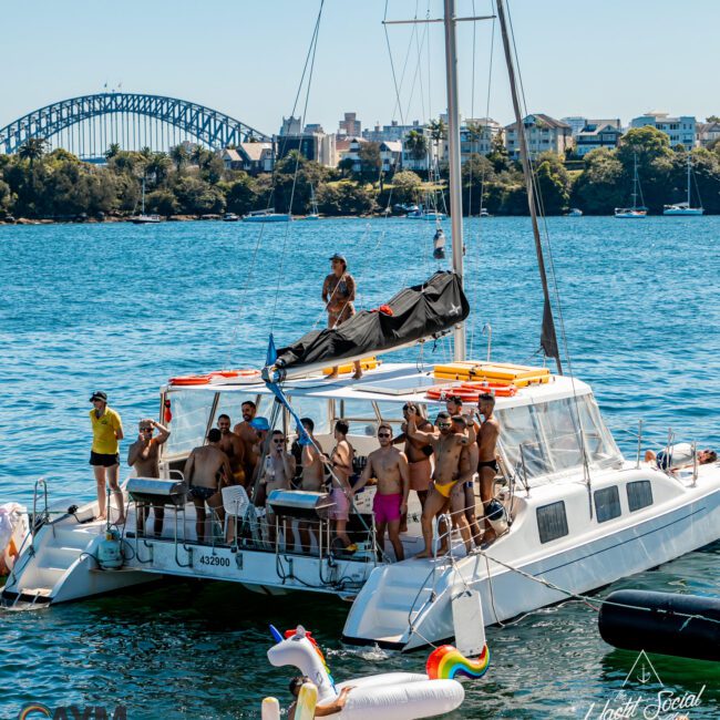 A group of people in swimsuits is enjoying a sunny day on a catamaran, with a city skyline and large bridge in the background. Some are on an inflatable unicorn in the water. Two logos are on the bottom corners of the image. Perfect for Boat Parties Sydney The Yacht Social Club.