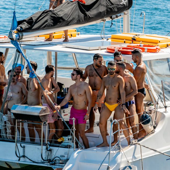 A group in swimwear gathers on a sailing boat, enjoying a sunny day on the water. Some stand on the deck, while one person is on the mast platform. Colorful inflatables float nearby. The overall mood appears lively and festive, capturing the essence of The Yacht Social Club Event Boat Charters.