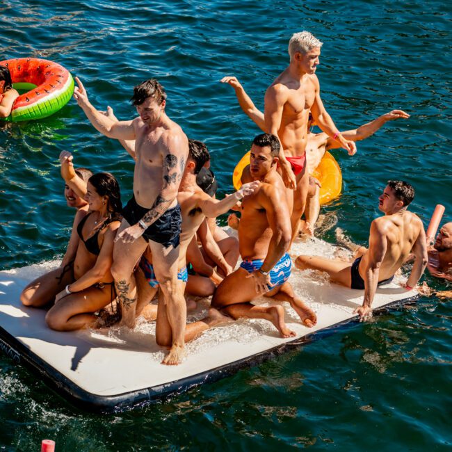 A group of people enjoying themselves on a large inflatable raft in the water. Some are dancing, while others are seated or lying down. They are surrounded by other colorful inflatables and are in a sunny outdoor setting, part of The Yacht Social Club Sydney Boat Hire event.