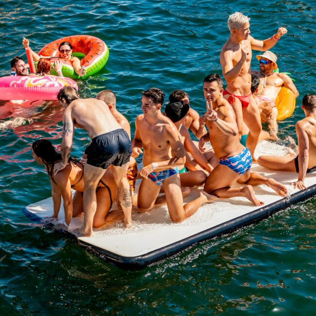 A group of people in swimsuits having fun on a floating platform in a body of water. Some are sitting or standing on the platform, while others are in the water nearby on inflatables. There is a social, party-like atmosphere under a sunny sky with The Yacht Social Club Sydney Boat Hire.