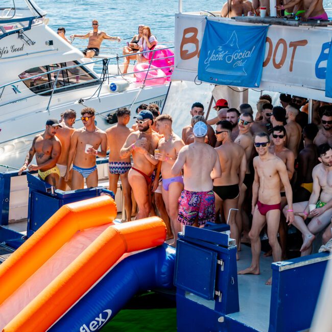 A lively scene on a boat with numerous people in swimwear, socializing and standing near an inflatable slide leading into the water. Another boat with people can be seen in the background on a sunny day. The atmosphere at this Yacht Social Club event is festive and vibrant, perfect for luxury yacht rentals in Sydney.