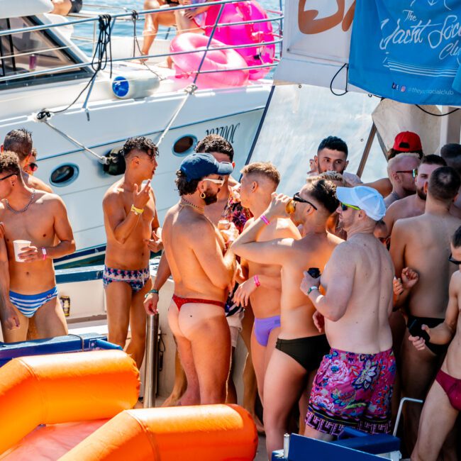 A group of men in swimwear are gathered on a boat in a celebratory atmosphere. Some hold drinks, while others engage in conversation. The Yacht Social Club Sydney Boat Hire has decorated the boat with banners, and the backdrop reveals another vessel on the water.