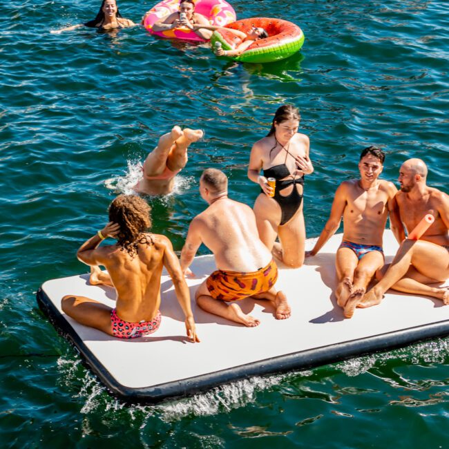 A group of people is enjoying a sunny day on the water. Several individuals are lounging on an inflatable platform, while others float nearby in colorful inner tubes. Everyone appears relaxed and happy. The vibrant blue water of Sydney Harbour surrounds them, with a boat from The Yacht Social Club partially visible in the background.