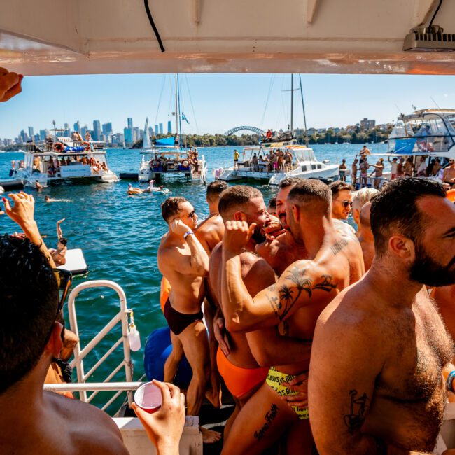 A lively yacht party on a sunny day, hosted by The Yacht Social Club Sydney Boat Hire, features shirtless men dancing closely together. Numerous boats are anchored nearby, with a stunning city skyline and a prominent bridge setting the perfect backdrop.