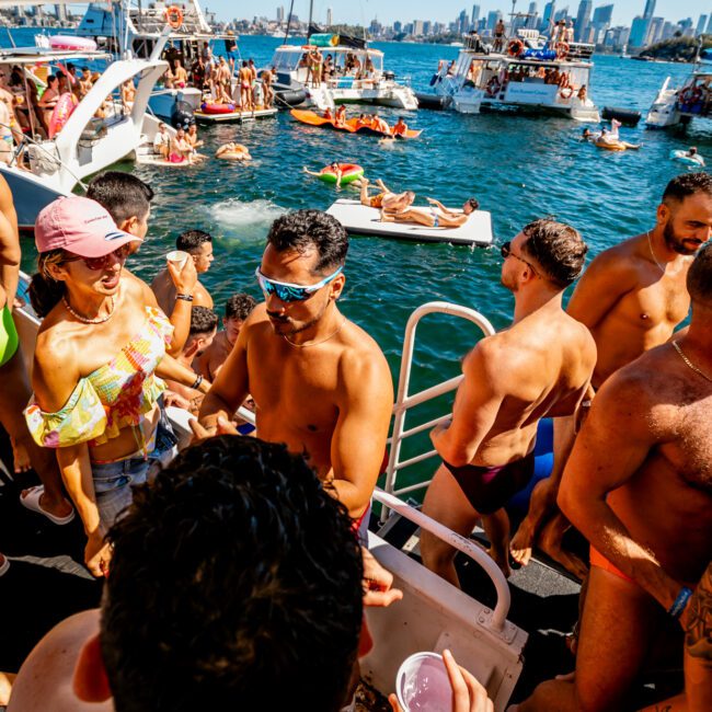 A group of people in swimwear are enjoying a sunny day on a boat at The Yacht Social Club Event. The water is filled with other boats and people swimming or lounging on floats. The scene is vibrant with a city skyline in the background. Some people are holding drinks, chatting, and dancing.