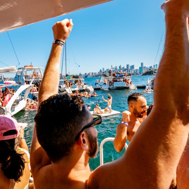 A vibrant scene of people partying on boats under clear blue skies, with a city skyline in the background. The crowd enjoys music and drinks, waving arms and dancing. A variety of boats from Sydney Harbour Boat Hire The Yacht Social Club are clustered together, creating a festive atmosphere.