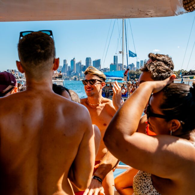 A group of people in swimwear are gathered on a boat during a sunny day. The background features a city skyline with tall buildings and boats on the water. The atmosphere appears lively and festive, with everyone enjoying the sunlight and socializing at The Yacht Social Club Sydney Boat Hire event.