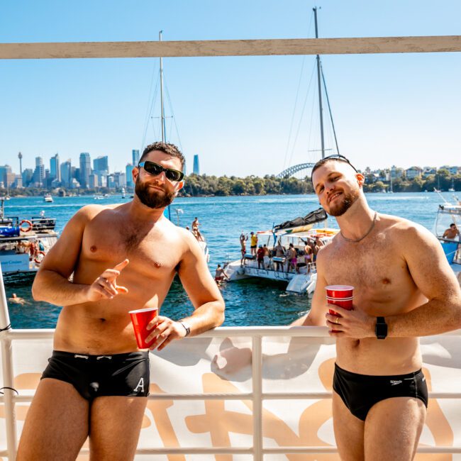 Two men wearing swim briefs stand on a boat, each holding a red cup. They are posing and smiling with the Sydney Harbour Bridge and city skyline in the background. Several other boats and people enjoy the sunny day, typical of Boat Parties Sydney with The Yacht Social Club. Logos are visible in image corners.