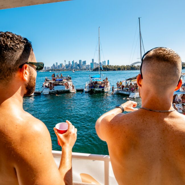 Two shirtless men are pictured from behind on a boat, holding drinks and looking out over the water. Multiple boats with people are seen in the distance against a backdrop of a city skyline. The image includes event logos in the bottom corners, capturing the essence of Sydney Harbour Boat Hire The Yacht Social Club.