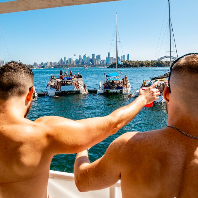 Two men on a boat enjoy a sunny day on the water, pointing towards other boats filled with people. The city skyline and a large bridge are visible in the background. The scene is lively, with clear blue skies and a festive atmosphere. Banners for Boat Parties Sydney The Yacht Social Club are seen.